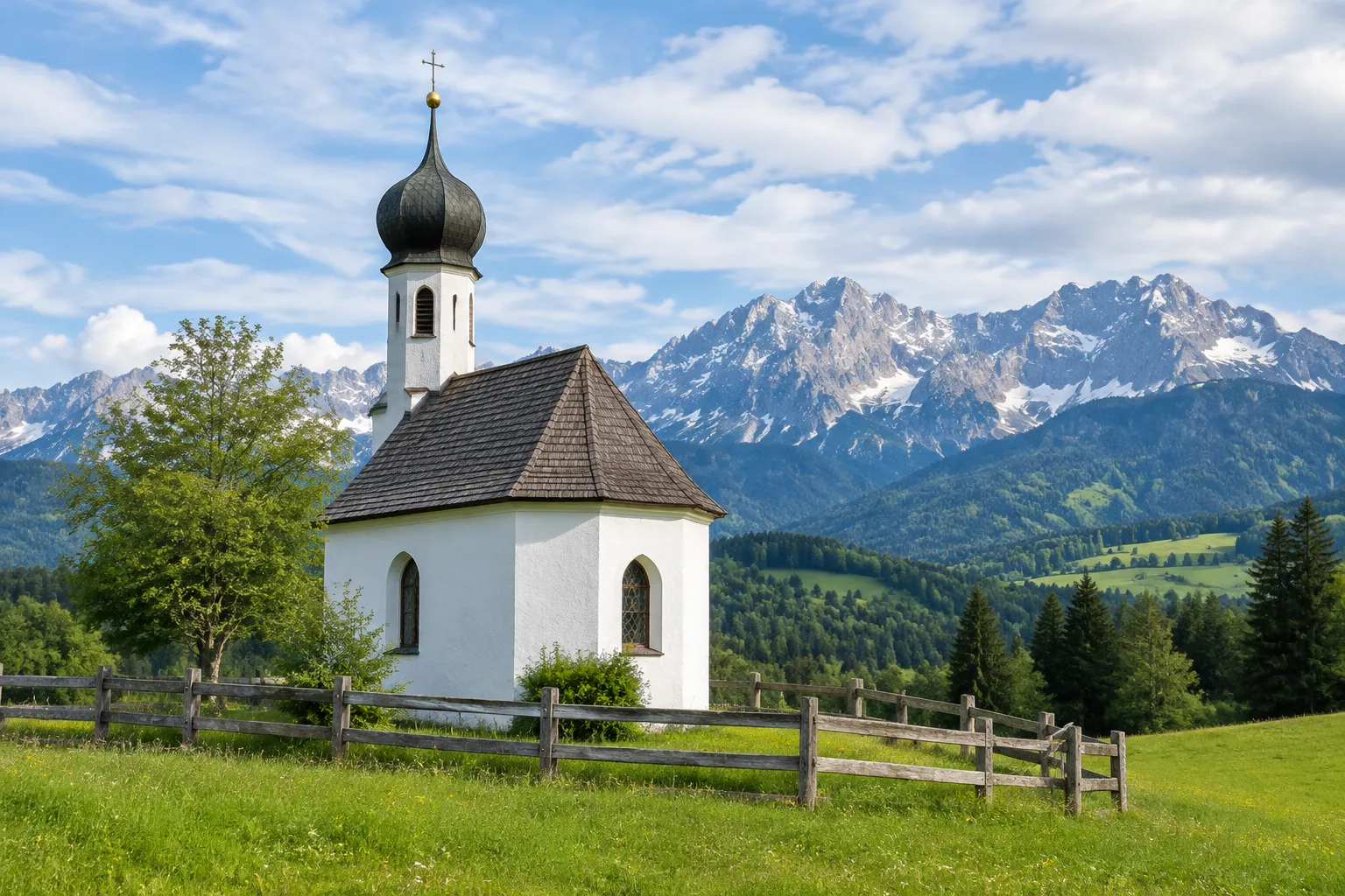 Weiße Kapelle mit Zwiebelturm auf einer Wiese im Alpenvorland, dahinter die Alpen